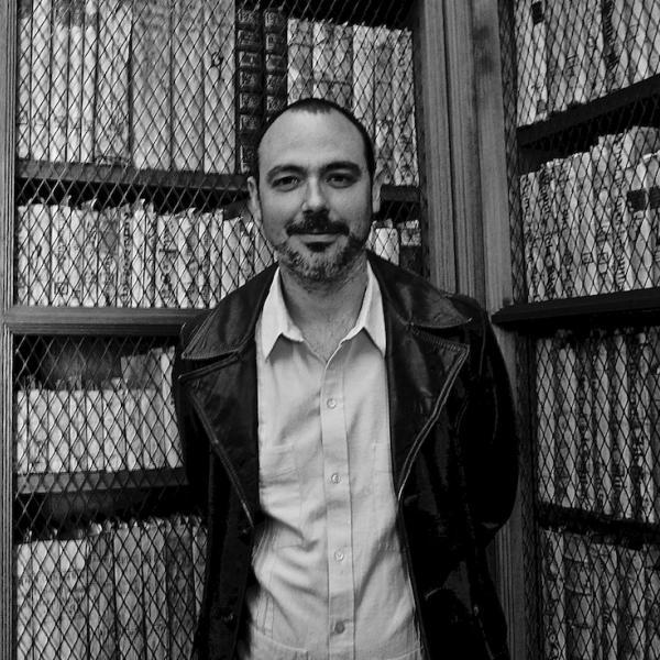 Black and white portrait of Miguel Martínez standing in front of a bookshelf