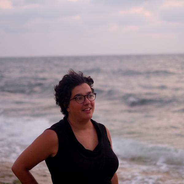 Portrait of Na'ama Rokem looking to the right in front of waves on a beach