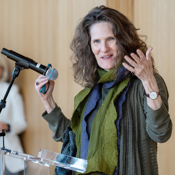 Portrait of Michèle Lowrie giving a presentation at a podium in front of a wood panel background